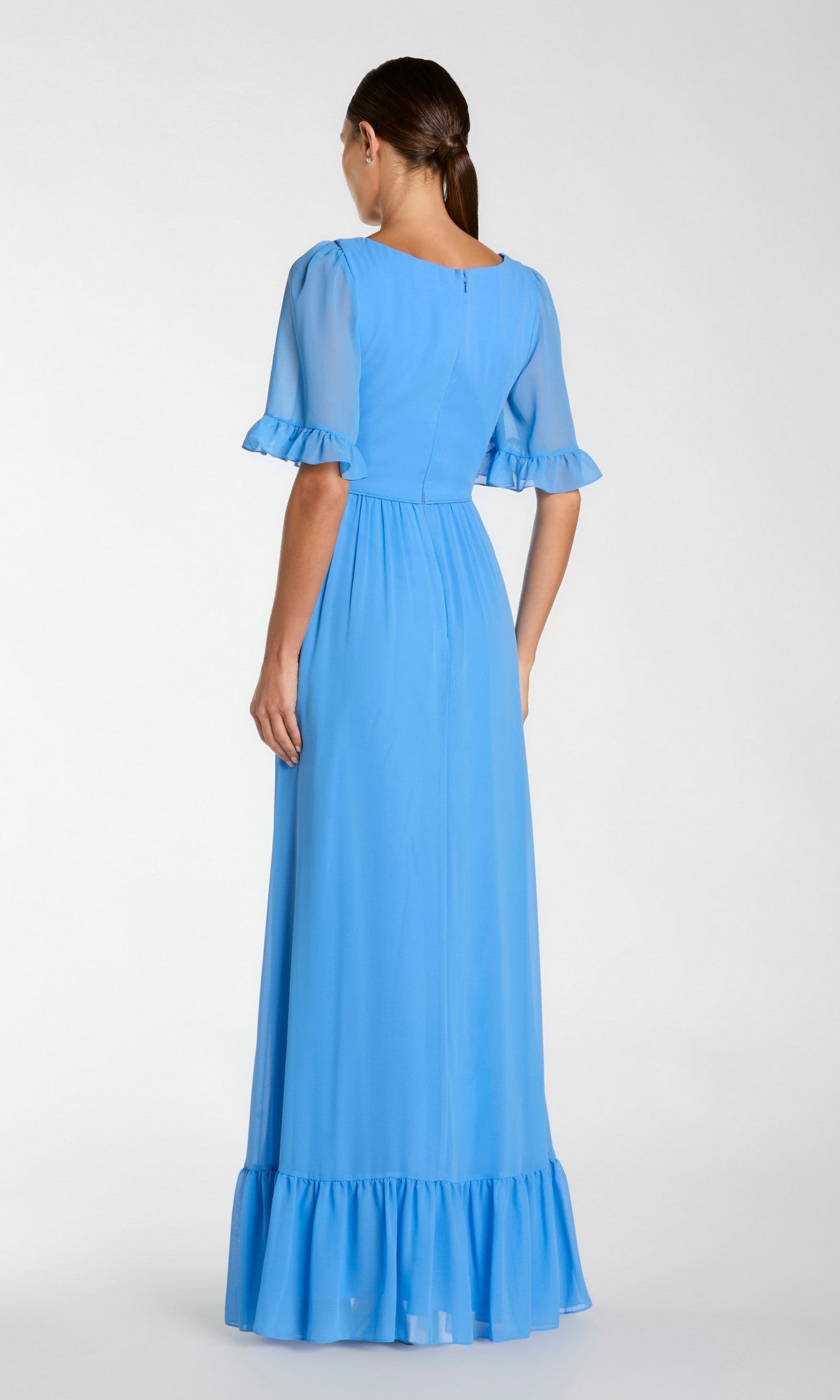 A woman stands with her back to the camera, wearing the Long Formal Dress 55867 by Mac Duggal— a light blue, short-sleeve, floor-length A-line dress with ruffle details on the sleeves and hem.